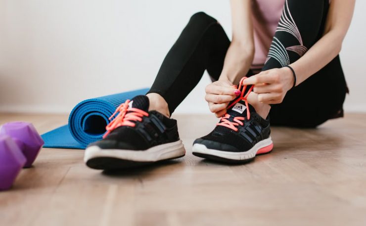 Crop sportswoman in sportswear tying sneakers while sitting on wooden floor with sport mat and dumbbells before exercising indoors